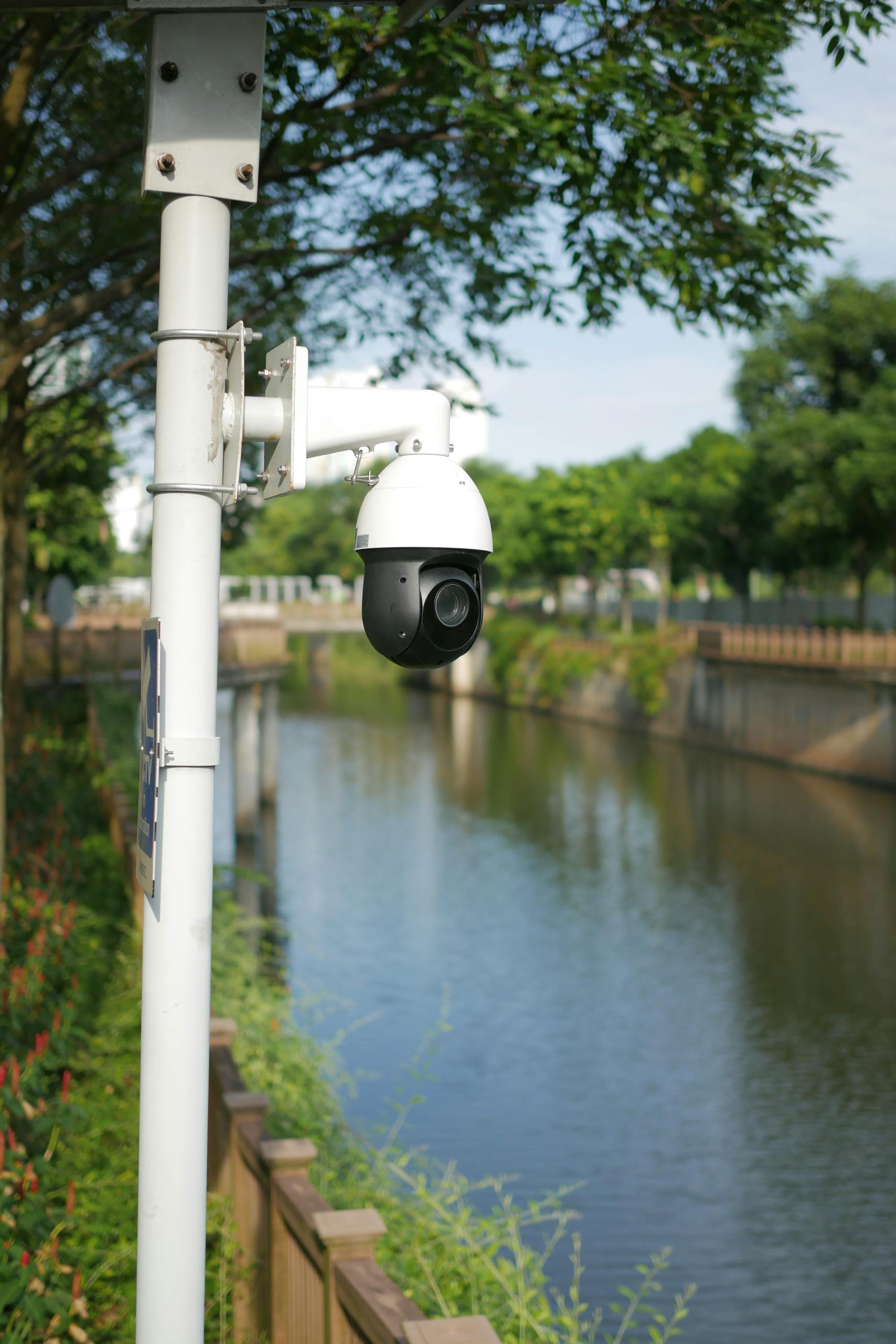 Close-up of a security camera near a tranquil river setting, surrounded by greenery.